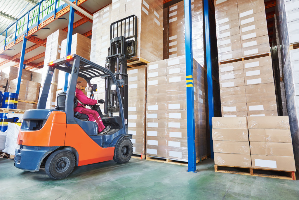 warehouse worker driver in uniform stacking cardboxes by forklift stacker loader
